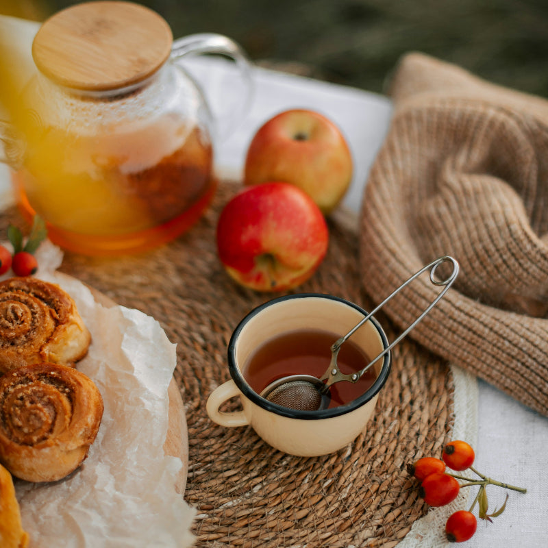 Tea set with a cup, teapot, and apples on a woven mat outdoors.
