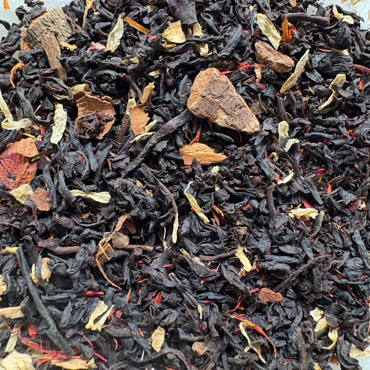 A detailed top-down view of the loose-leaf Cocoa Gingerbread black tea blend, featuring dark black tea leaves mixed with chunky pieces of cinnamon bark, star anise fragments, dried ginger, light cocoa nibs, and scattered orange safflower petals.
