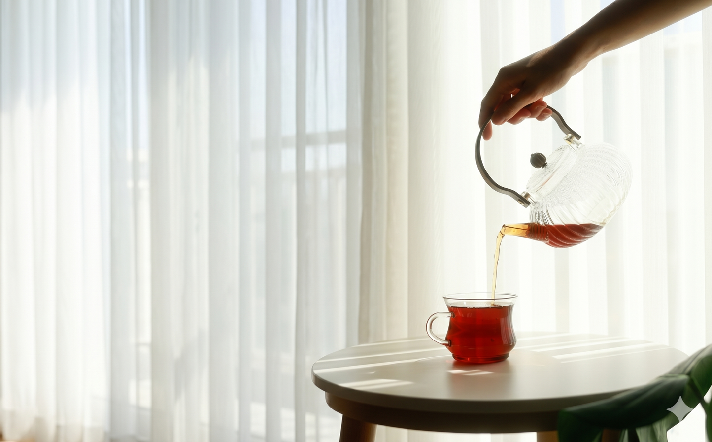 Person pouring tea from a glass teapot into a cup on a table with a blurred background