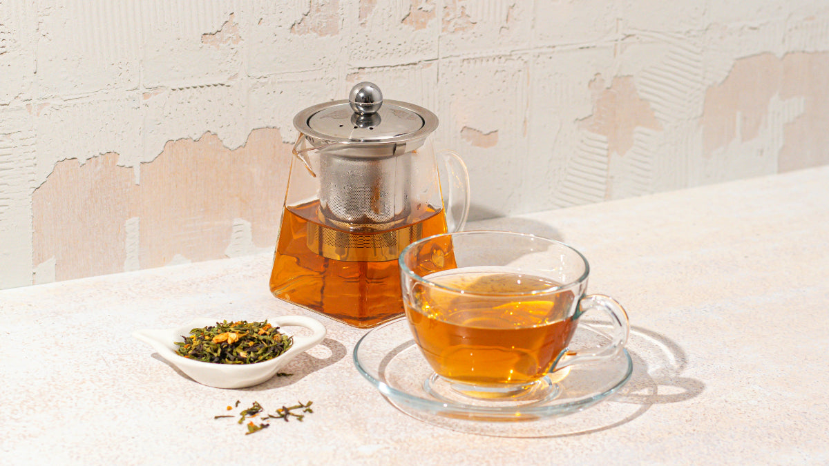 Tea set with a glass teapot, cup, and saucer on a textured surface.