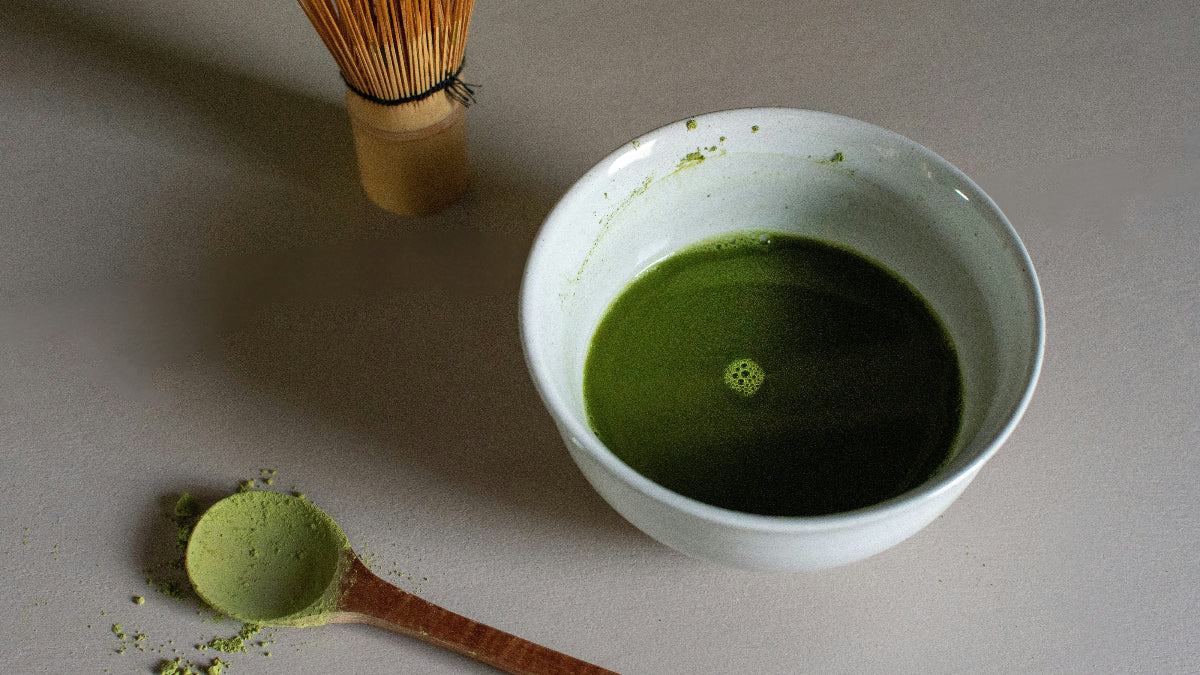 Matcha green tea preparation with a bowl of matcha, whisk, and spoon on a light surface.