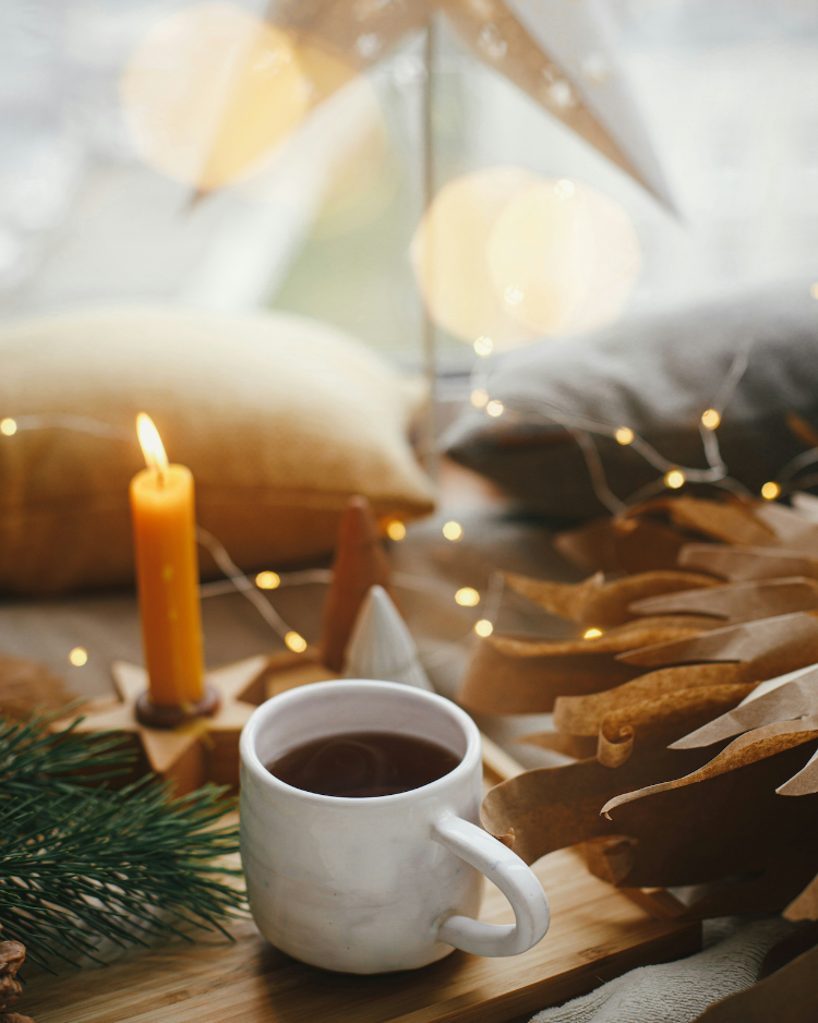 White mug with hot tea on a wooden table with a candle and decorative pillows in the background.