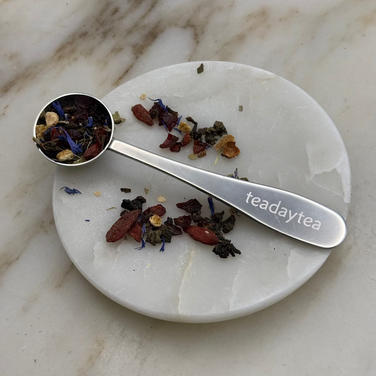 Tea spoon with tea leaves on a marble surface, featuring 'teadaytea' branding.