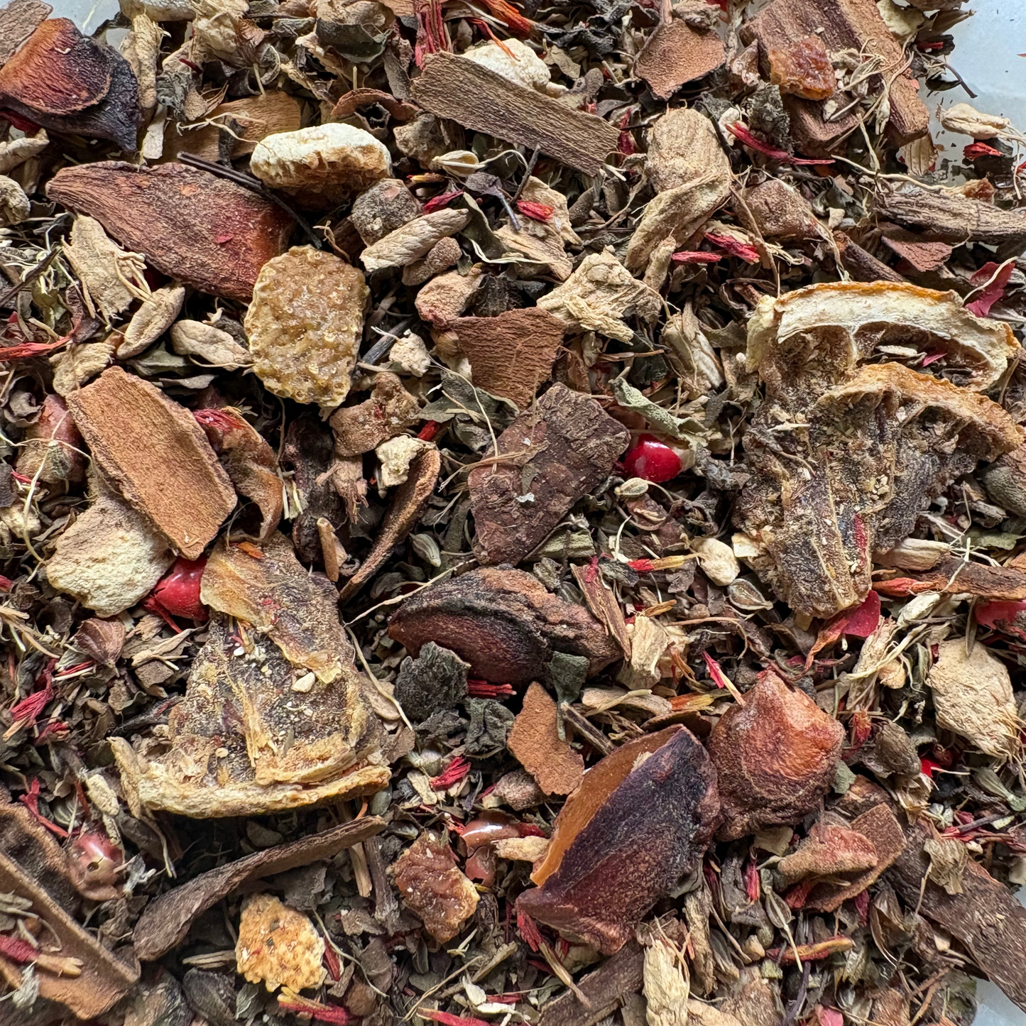 A vibrant loose-leaf tea blend of Zesty Tulsi featuring a rich mixture of textured green tulsi leaves, fragments of dried ginger root, pieces of star anise, and bright, sun-dried citrus peels piled on a white surface.