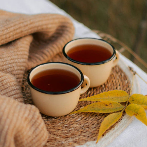 Two cups of tea on a woven mat with a blanket and autumn leaves.