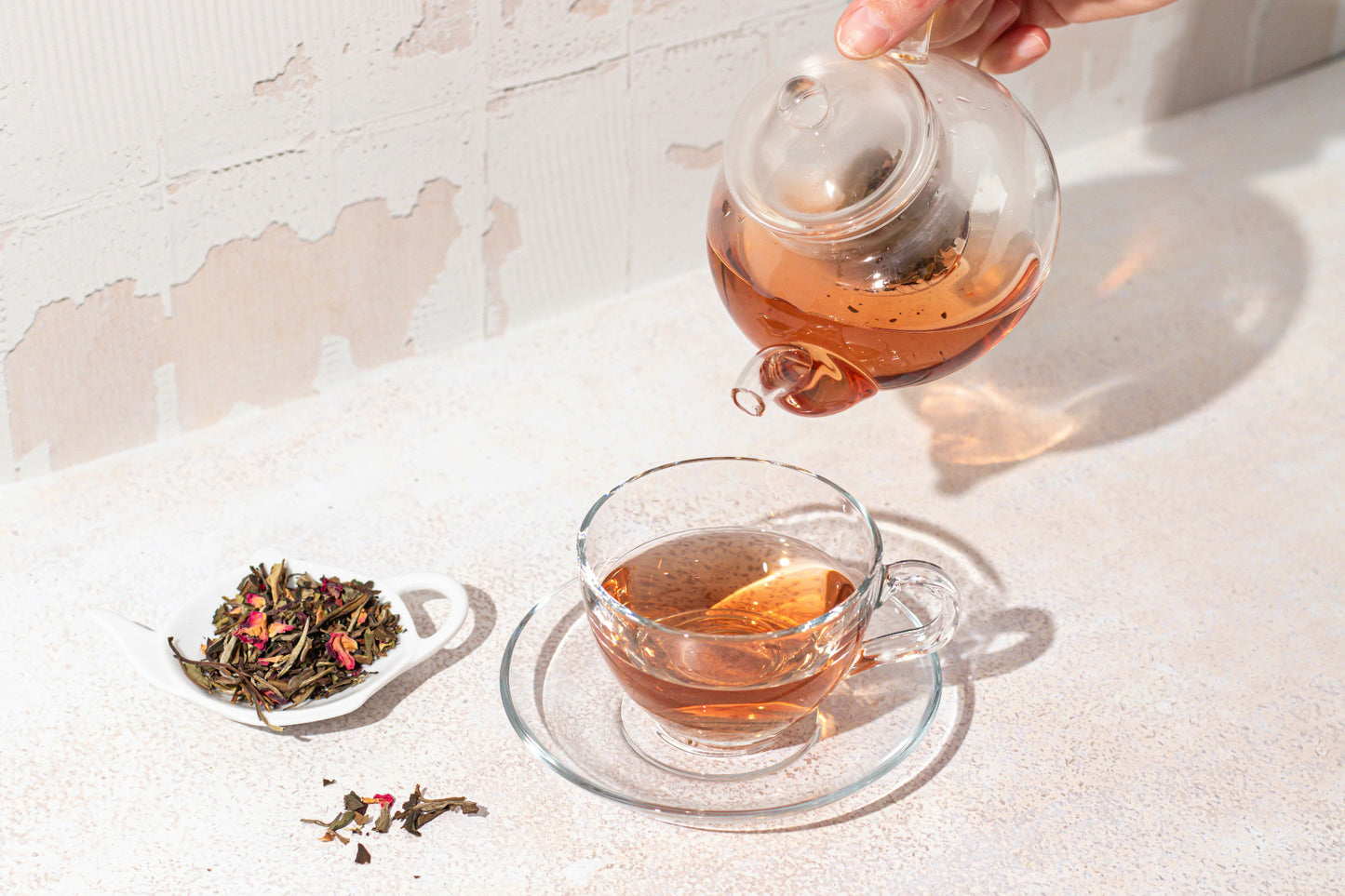 Tea being poured from a glass teapot into a glass cup on a light surface.
