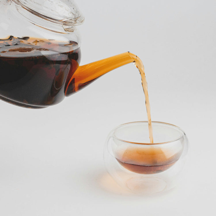 Glass teapot pouring black tea into a glass cup on a light gray background