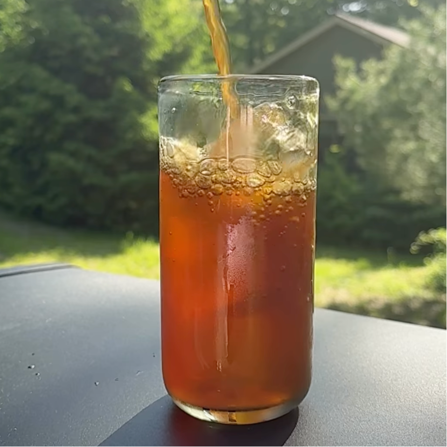 A rich amber iced tea being poured from a glass pitcher into a clear, condensation-covered glass filled with ice, capturing a refreshing glass of Candied Ginger Peach rooibos tea outdoors.