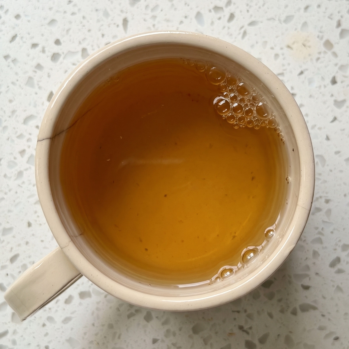A top-down view of a ceramic mug filled with steeped, golden-amber Tranquility herbal tea with small bubbles on the surface, sitting on a light, speckled countertop.