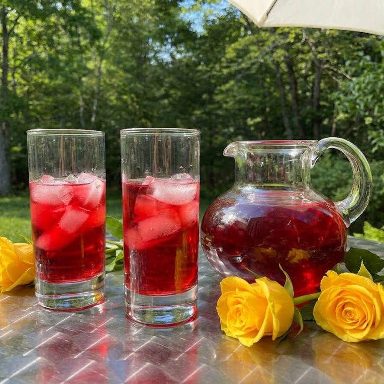 Two clear glasses filled with a vibrant red iced tea and large ice cubes sit on a metal outdoor table next to a matching glass pitcher. Bright yellow roses rest on the table in the foreground against a soft-focus background of lush green trees and a white patio umbrella under bright daylight.